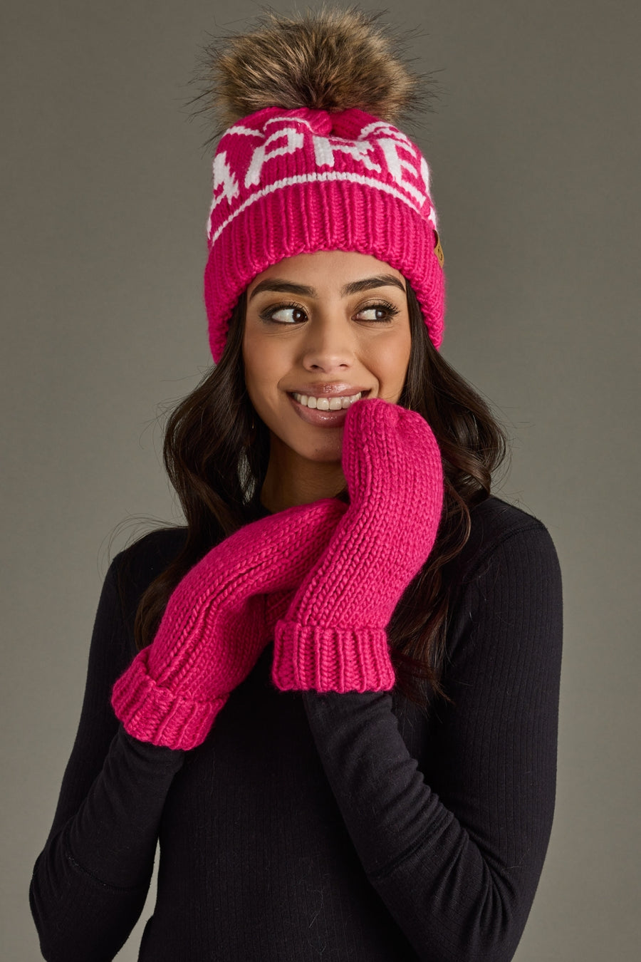 Person wearing a pink knit hat and mittens with a brand logo on a gray background