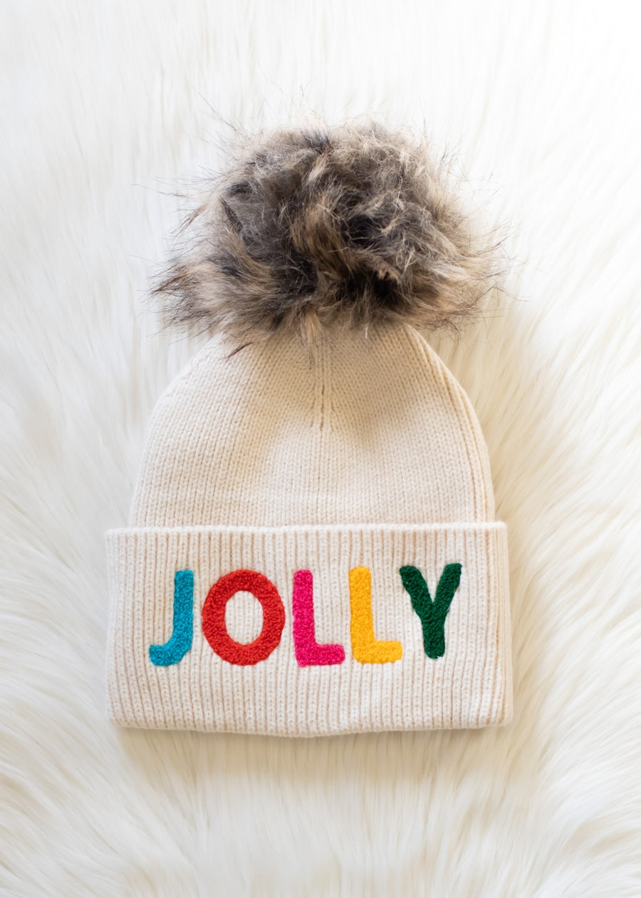 Beige knit beanie with colorful 'JOLLY' text and fur pom-pom on a white background