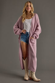 Woman wearing a long pink cardigan over a white top and denim shorts, standing against a gray background.