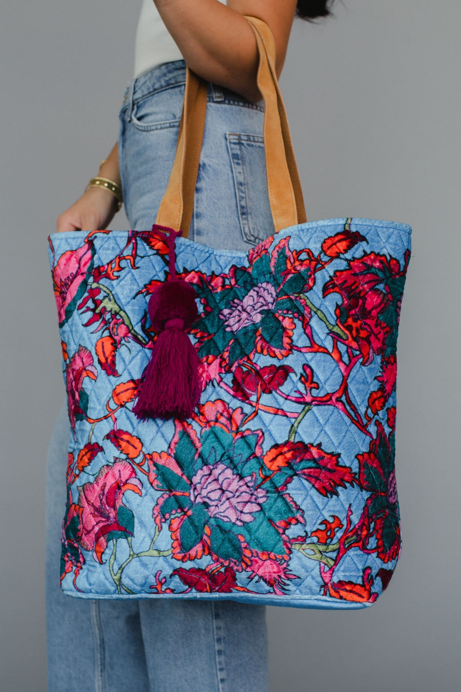 Floral patterned tote bag with a tassel held by a person wearing jeans against a gray background
