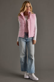 Woman wearing a pink puffer vest, white shirt, blue jeans, and white sneakers on a gray background