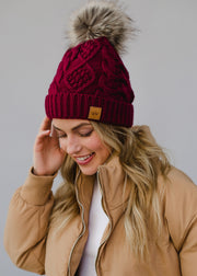 Woman wearing a maroon knit beanie with a fur pom-pom and a beige jacket on a gray background