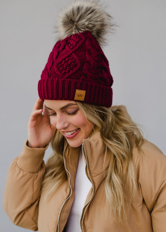 Woman wearing a maroon knit beanie with a fur pom-pom and a beige jacket on a gray background