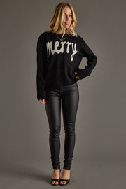 A woman with long blonde hair stands against a plain background, wearing the Merry Sweater - Black from Panache Apparel Collection, paired with leather-look leggings and open-toe heels. She smiles slightly.