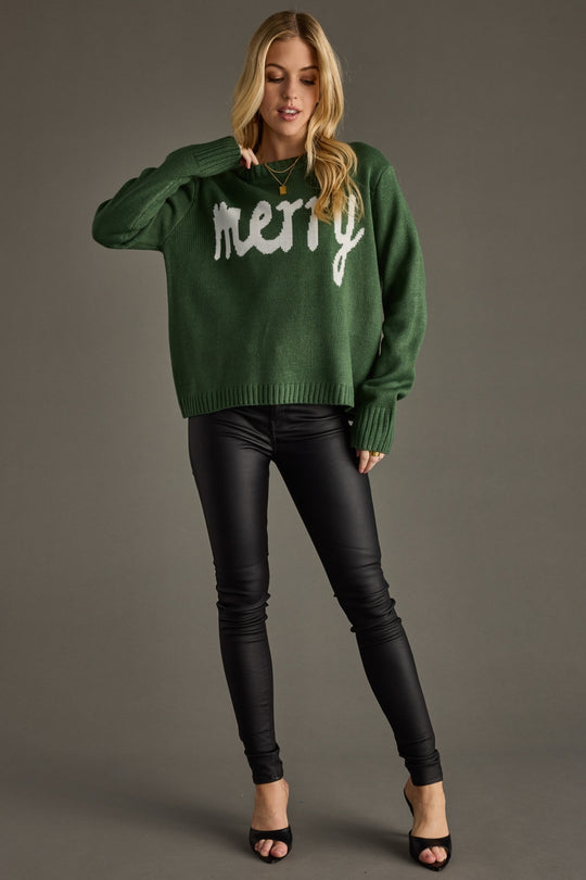 Woman wearing a green sweater with 'merry' text and black pants on a gray background