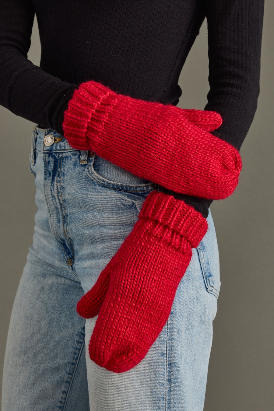 Red knitted mittens worn with a black long-sleeve shirt and blue jeans on a neutral background