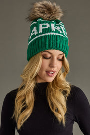 Green knit beanie with fur pom-pom worn by a person against a gray background