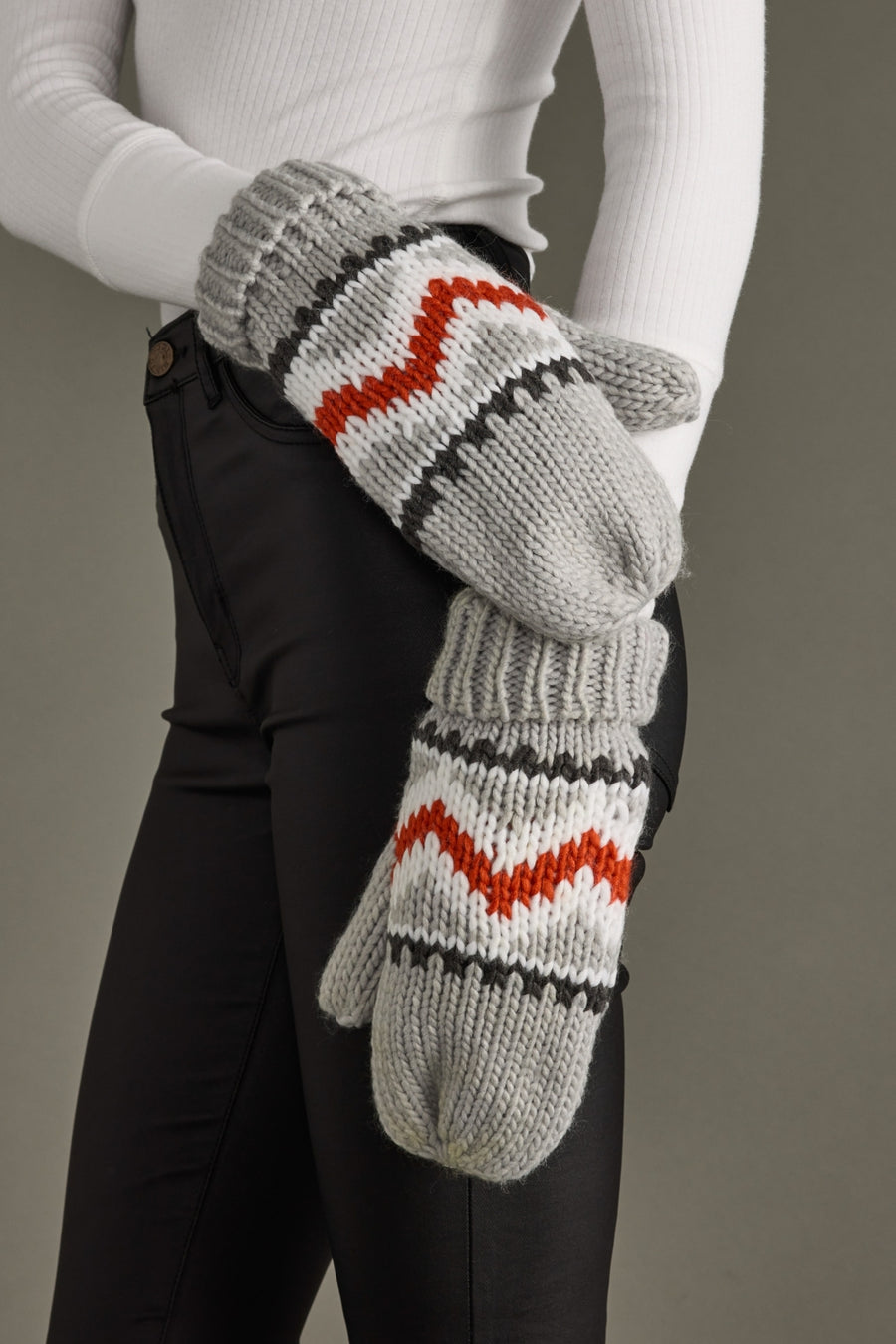 Gray knitted mittens with red and black pattern worn by a person against a neutral background