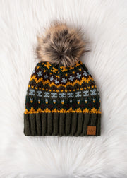 The Reyna Pom Hat by Panache Apparel Collection features a colorful knit design, large faux fur pom-pom, and cozy fleece lining for extra warmth, shown on a white, fluffy surface.