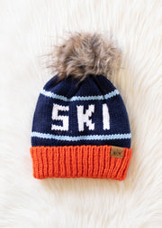 Knitted beanie with 'SKI' text and fur pom-pom on a light wooden surface
