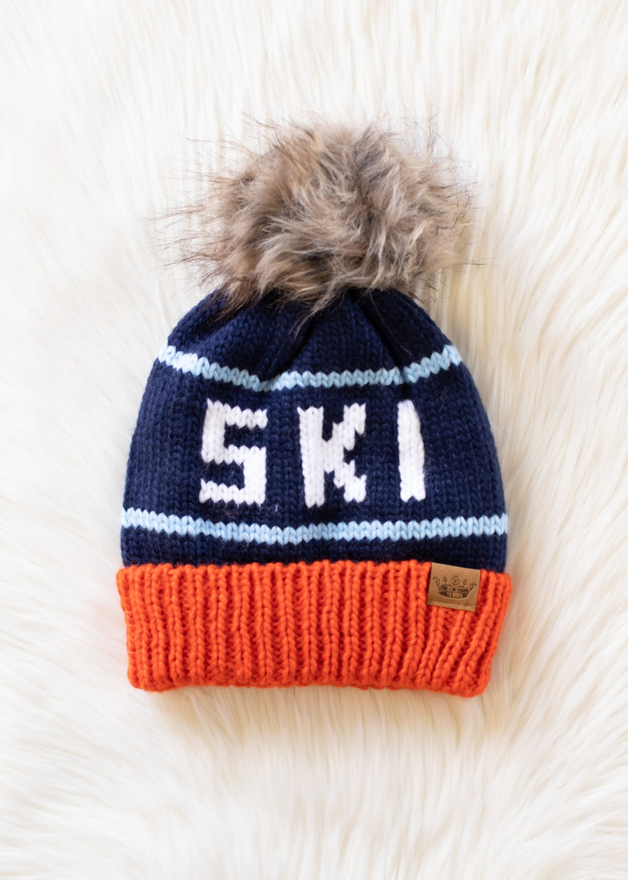 Knitted beanie with 'SKI' text and fur pom-pom on a light wooden surface