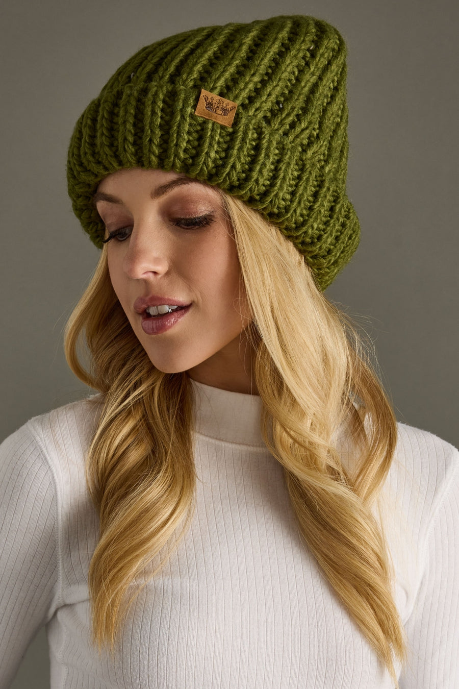 Woman wearing a green knitted beanie with a brand logo against a gray background