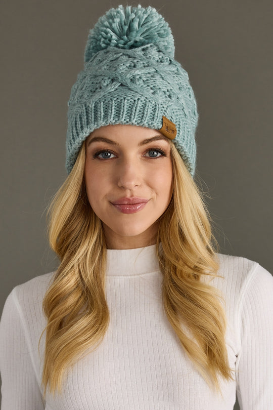 Woman wearing a knit teal beanie with a pom-pom against a gray background