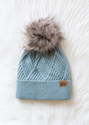 Blue knit beanie with fur pom-pom on a white textured background