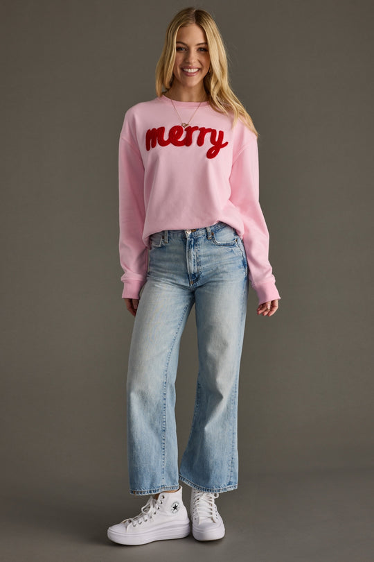 Woman wearing a pink sweatshirt with 'merry' text, blue jeans, and white sneakers on a gray background