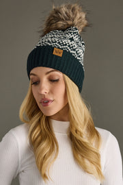 Woman wearing a dark teal and white patterned beanie with a fluffy pom-pom on a gray background