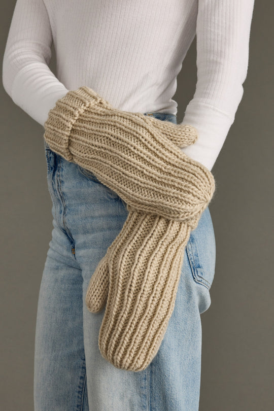 Beige knitted mittens worn with a white long-sleeve shirt and blue jeans against a neutral background