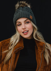 A woman with long blonde hair looks directly at the camera with a soft, relaxed expression, wearing a dark gray cable-knit beanie with a faux-fur pom-pom and a small leather logo patch on the front. She’s styled in a black top layered under a warm brown jacket, set against a clean, dark background that emphasizes her natural makeup and cozy fall–winter aesthetic.