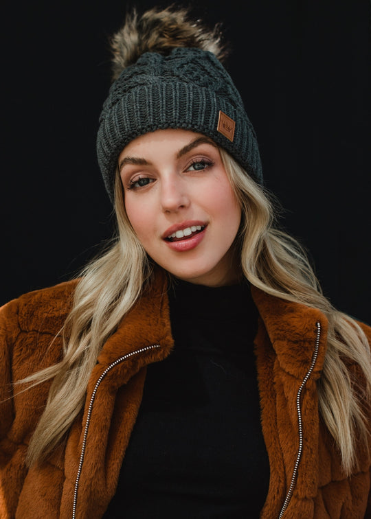 A woman with long blonde hair looks directly at the camera with a soft, relaxed expression, wearing a dark gray cable-knit beanie with a faux-fur pom-pom and a small leather logo patch on the front. She’s styled in a black top layered under a warm brown jacket, set against a clean, dark background that emphasizes her natural makeup and cozy fall–winter aesthetic.