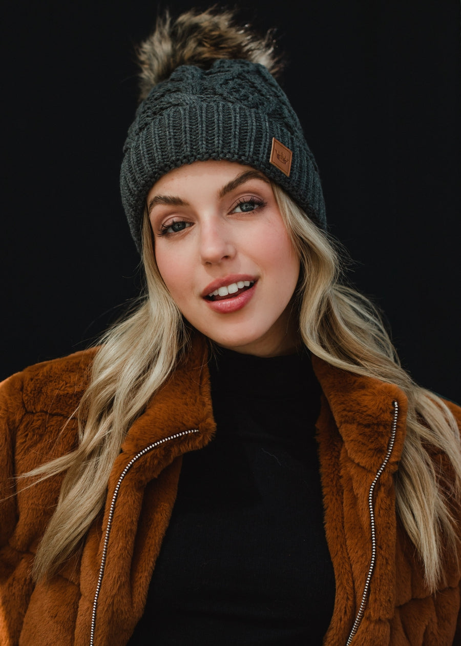 A woman with long blonde hair looks directly at the camera with a soft, relaxed expression, wearing a dark gray cable-knit beanie with a faux-fur pom-pom and a small leather logo patch on the front. She’s styled in a black top layered under a warm brown jacket, set against a clean, dark background that emphasizes her natural makeup and cozy fall–winter aesthetic.