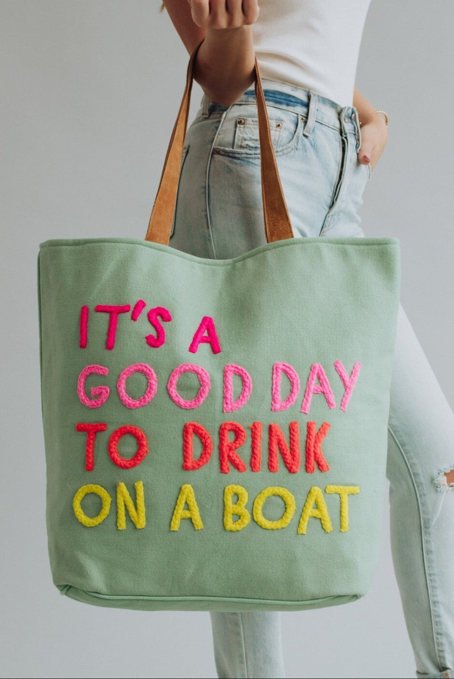 A person holds the Mint Drink On A Boat Tote from Panache Apparel Collection, featuring leather suede straps and colorful lettering that says, “IT’S A GOOD DAY TO DRINK ON A BOAT.” They are dressed in light blue jeans and a white top.