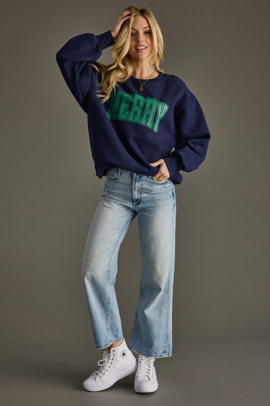 Woman wearing a navy sweatshirt with green text, light blue jeans, and white sneakers on a gray background
