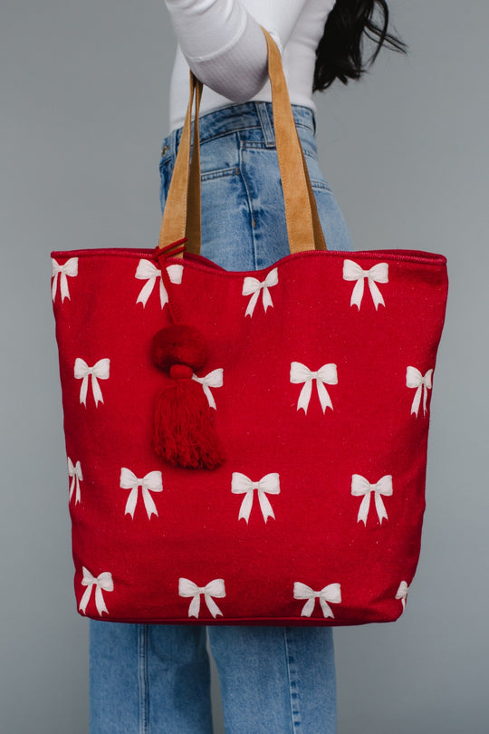 Red tote bag with white bow pattern held by a person wearing blue jeans against a gray background