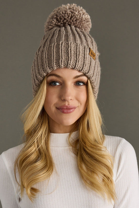 Woman wearing a taupe knit beanie with a pom-pom on a gray background