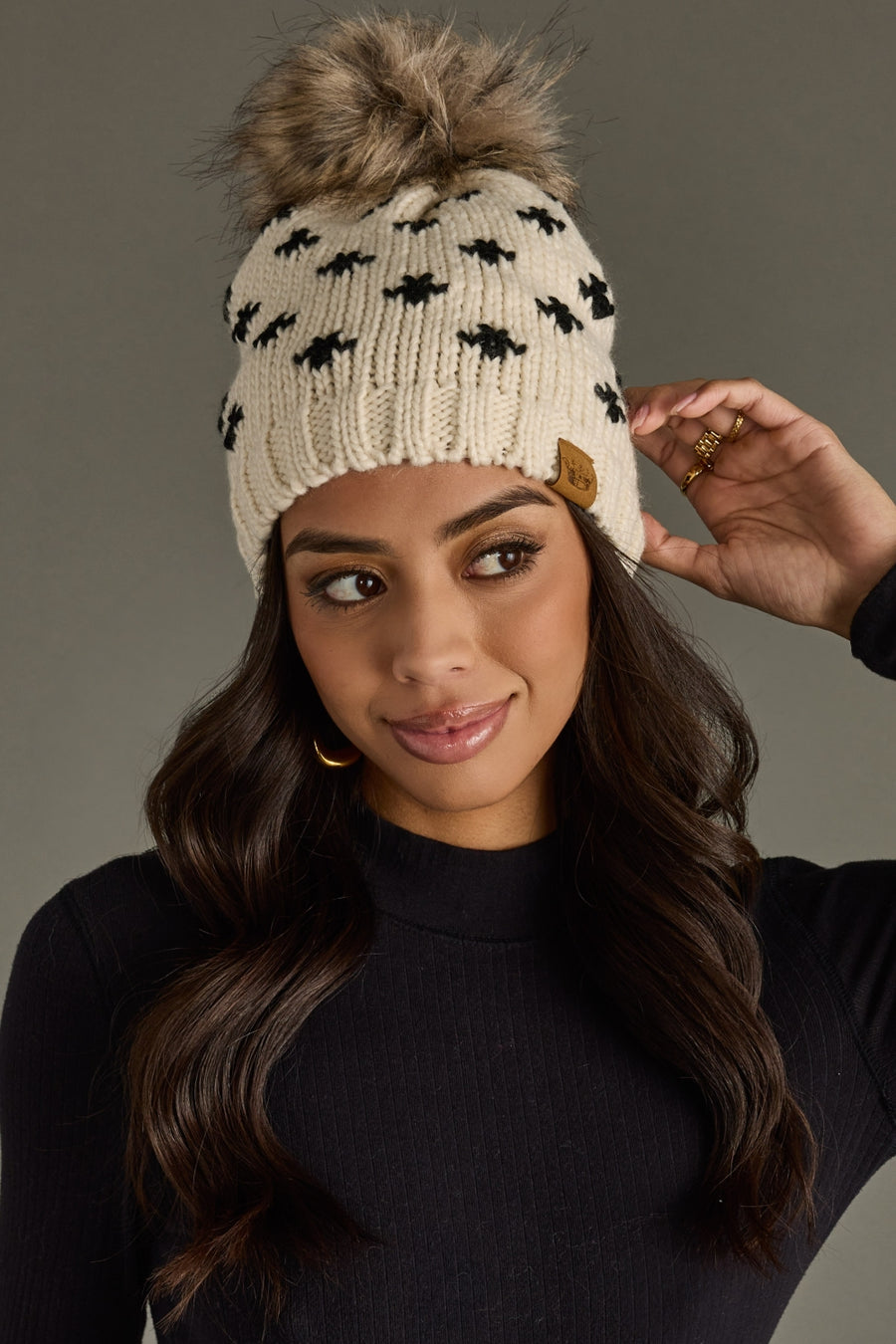 Woman wearing a cream beanie with black patterns and a fur pom-pom against a gray background