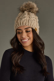 Woman wearing a beige knitted beanie with a pom-pom against a gray background