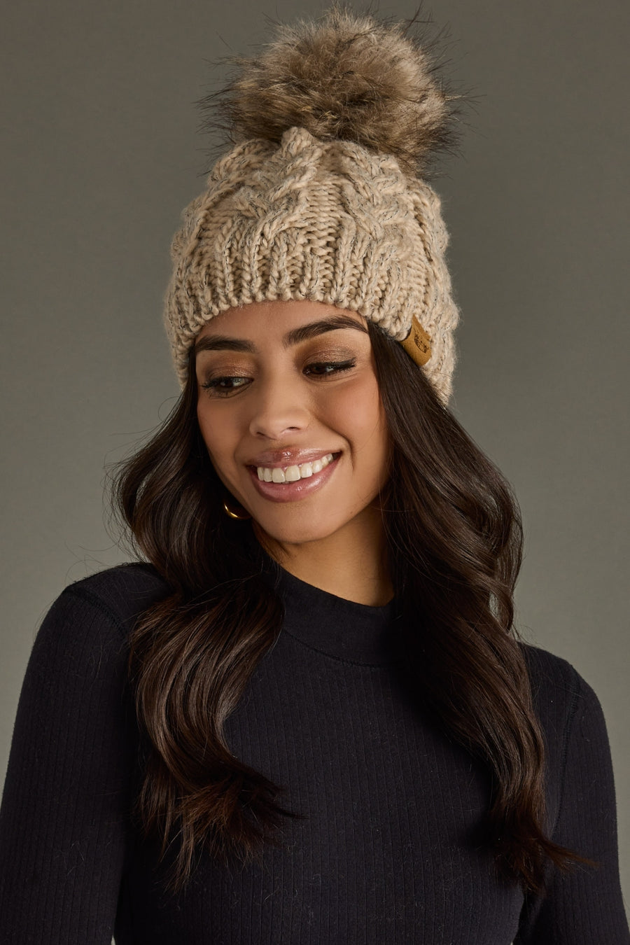 Woman wearing a beige knitted beanie with a pom-pom against a gray background