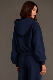 A woman with long brown hair wears the Dark Navy Panache Cropped Hooded Sweatshirt from the Panache Apparel Collection, paired with matching pants. She stands mostly facing away from the camera, glancing over her shoulder, against a plain gray background.