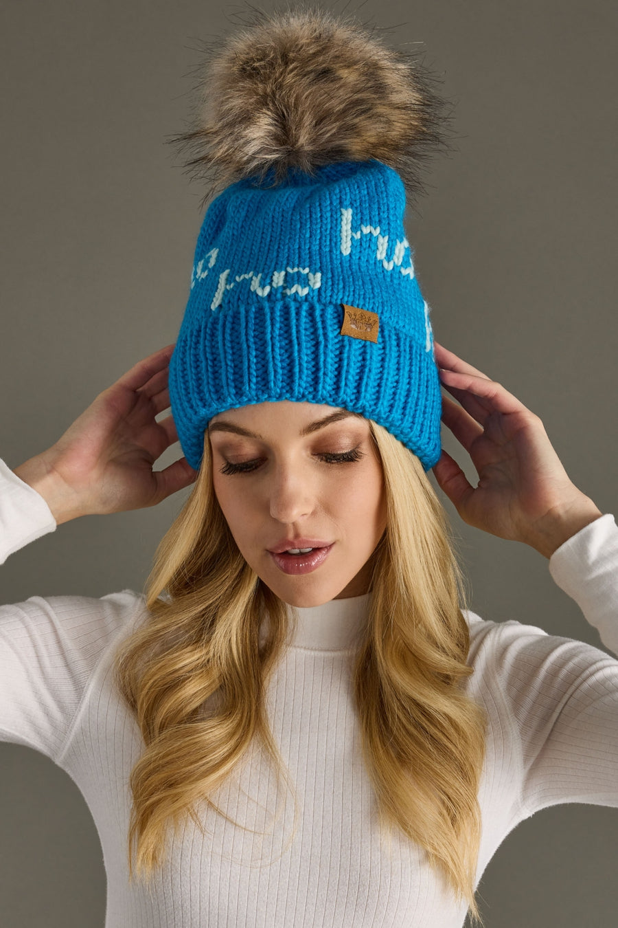 Woman wearing a blue knit beanie with a fur pom-pom against a gray background