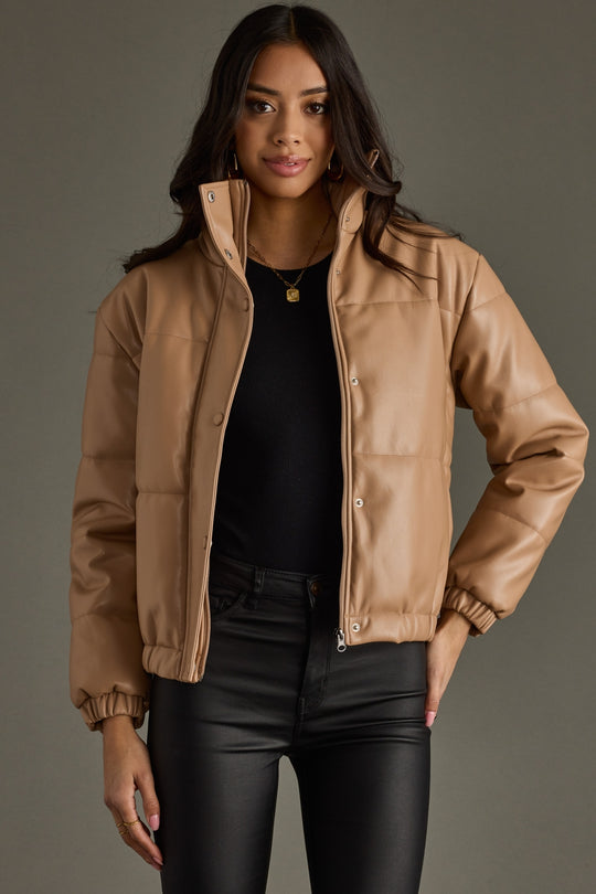 Woman wearing a tan jacket over a black top and pants on a gray background