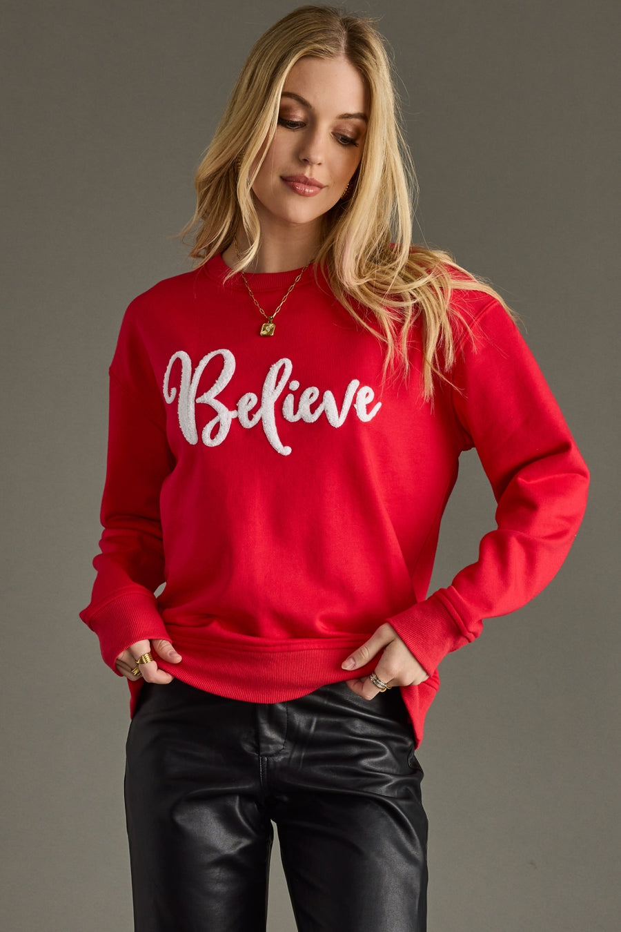 A woman models the Panache Apparel Collection's Believe Sweatshirt - Red, styled with black leather pants and gold jewelry. The relaxed fit sweatshirt features "Believe" in bold white script against a plain gray background.