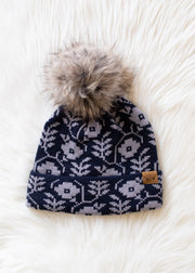 Navy blue knit beanie with white floral pattern and fur pom-pom on a white fluffy surface