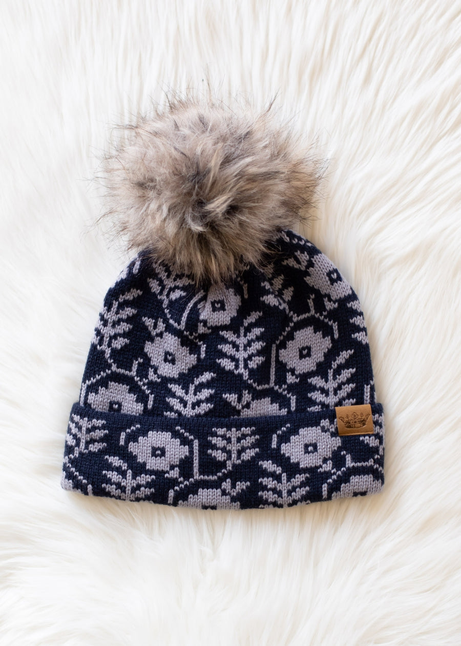 Navy blue knit beanie with white floral pattern and fur pom-pom on a white fluffy surface
