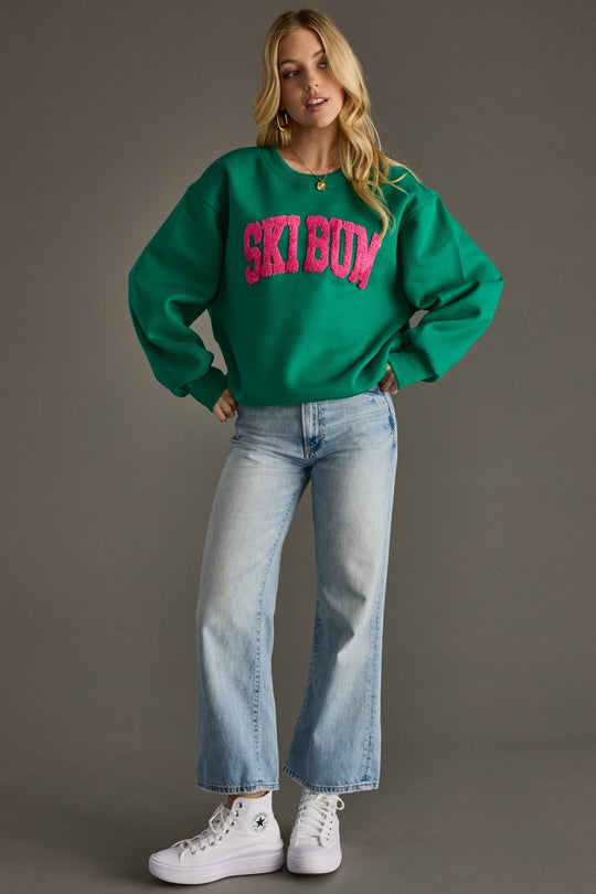 Woman wearing a green sweatshirt with 'SKI BUM' text, blue jeans, and white sneakers on a gray background
