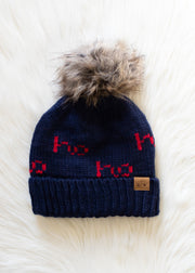 The Ho Ho Ho Pom Hat - Navy by Panache Apparel Collection features bold red "Ho Ho Ho" lettering, a cozy fleece-lined interior, and a brown faux fur pom-pom, shown on a white fluffy surface.