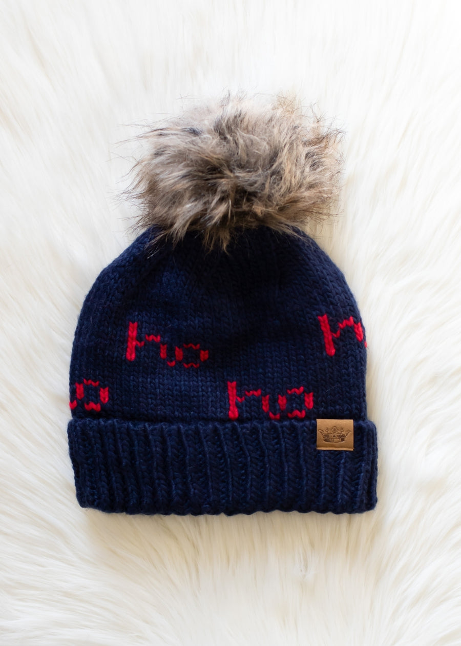 The Ho Ho Ho Pom Hat - Navy by Panache Apparel Collection features bold red "Ho Ho Ho" lettering, a cozy fleece-lined interior, and a brown faux fur pom-pom, shown on a white fluffy surface.