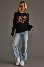 Woman wearing a black sweater with 'GAME DAY' text, blue jeans, and white sneakers on a gray background