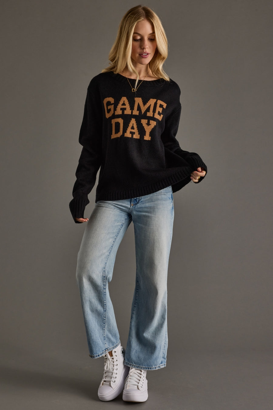 Woman wearing a black sweater with 'GAME DAY' text, blue jeans, and white sneakers on a gray background