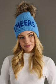 Woman wearing a blue beanie with 'CHEERS' text and a fur pom-pom on a gray background