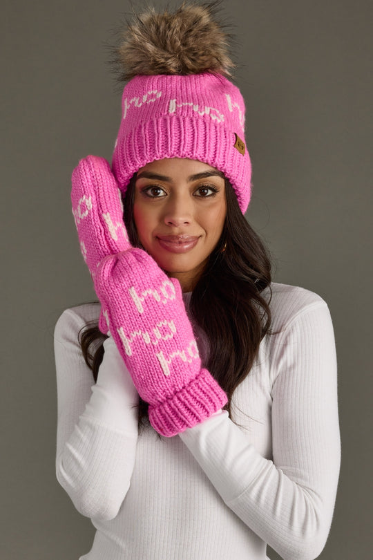 Woman wearing a pink knit beanie and gloves with a brown pom-pom against a gray background