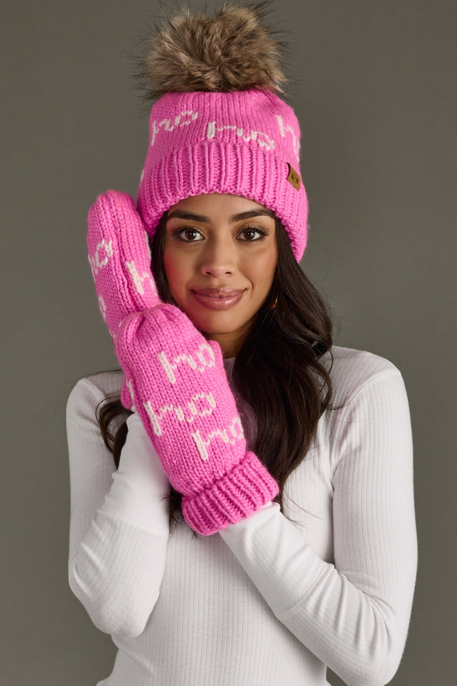 Woman wearing a pink knit beanie and gloves with a brown pom-pom against a gray background