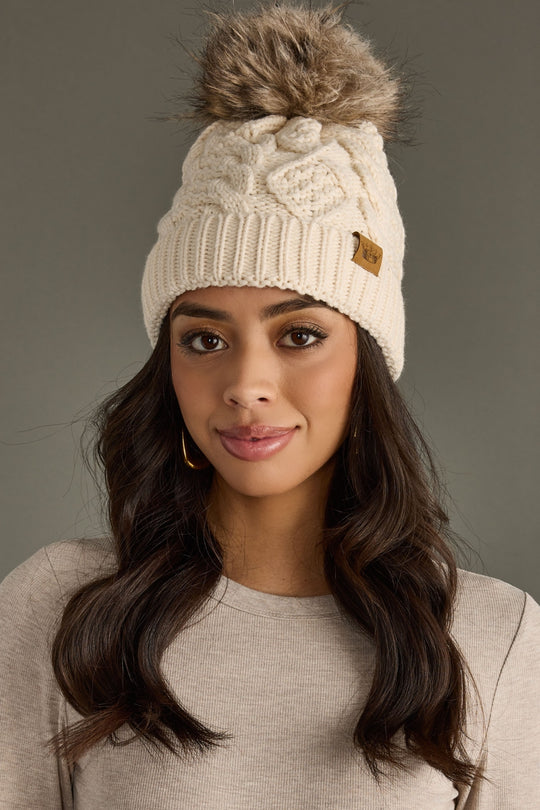 Woman wearing a cream knit beanie with a fur pom-pom against a gray background