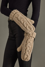 Beige knit mittens held by a person wearing black pants against a gray background