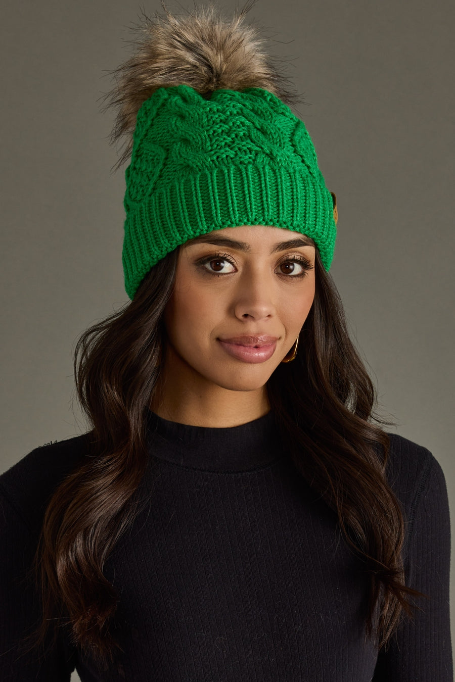 Woman wearing a green knitted beanie with a fur pom-pom against a gray background