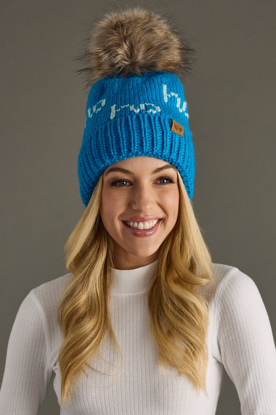 Woman wearing a blue knit beanie with a fur pom-pom on a gray background