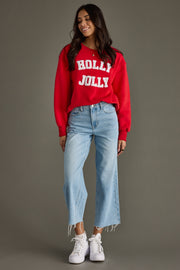 Woman wearing a red 'Holly Jolly' sweater with light blue jeans and white sneakers on a gray background